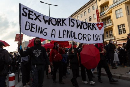 Des manifestants à Berlin habillés en rouge et en noir avec une pancarte avec écrit dessus "Sex work is work *cœur avec parapluie dedans* Being a landlord isn't" - traduction : "Les métiers du sexe sont des métiers, Être rentier n'en est pas un"