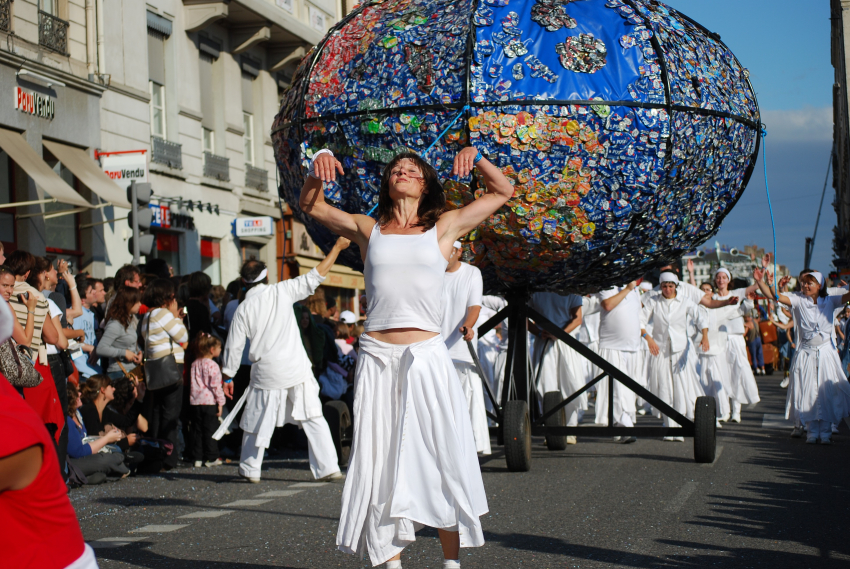 Annick Charlot, habillée en blanc dansant devant un char représentant la planète et fabriqué en canettes de bière et de soda utilisées.